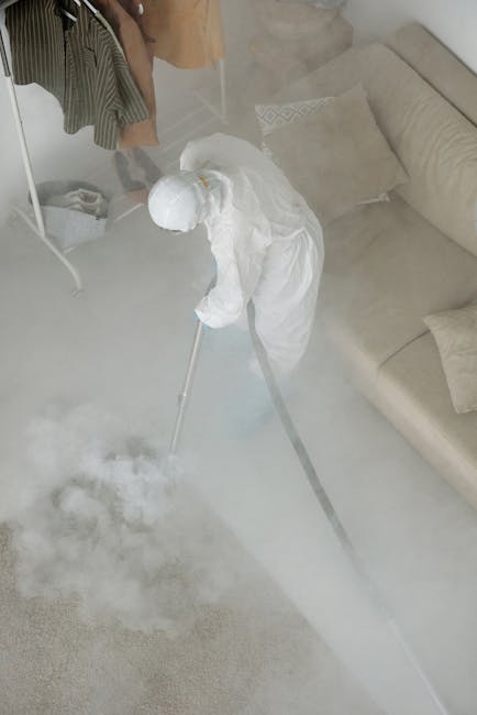 A person dressed in protective white clothing and a hood is performing disinfectant fogging or sanitisation in a living room, using a fogging machine that emits a cloud of disinfectant mist onto a lig