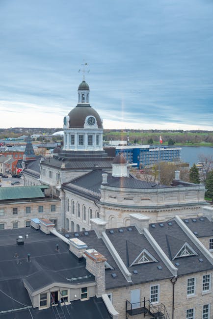A high-angle view of a historic cityscape featuring a large, ornate building with a prominent clock tower and a cupola. The building has a white facade with classical architectural details, contrastin