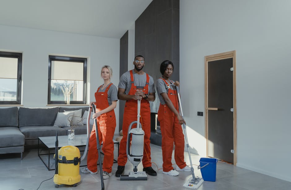 Two professional cleaning specialists, dressed in red overalls and grey shirts, are performing surface cleaning and deep cleaning on a modern living room's light wooden floor with vacuum cleaners. The