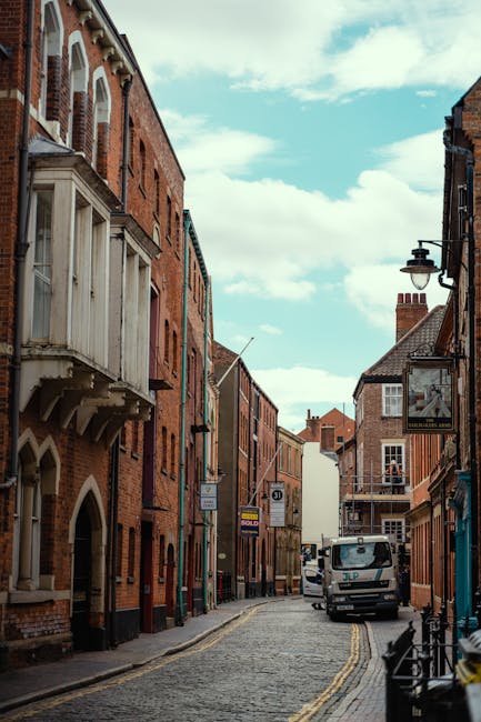 Photograph of a narrow cobblestone street on Kingston High Street, featuring historic brick buildings with varied architectural styles, including arched windows and decorative stonework. The street is lined with traditional street lamps and small shop signs, with a white commercial van parked along the sidewalk. The scene is illuminated by natural daylight, with a partly cloudy sky overhead. The setting showcases a quaint, well-maintained urban area in Kingston, emphasizing the aesthetic environment in which Kingston Carpet Cleaners provides its professional cleaning and surface sanitisation services for residential and commercial properties.