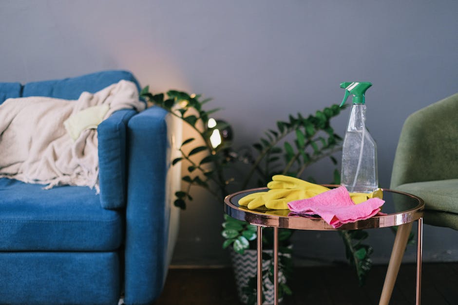 A living room scene featuring a blue upholstered sofa with a cream-colored throw blanket draped over its armrest, situated against a grey wall with a large green leafy plant nearby. Next to the sofa is a small, round, metallic side table with a polished rose gold finish, holding yellow rubber gloves, a pink microfiber cloth, and a transparent spray bottle filled with cleaning solution. The room is well-lit, highlighting the clean and tidy appearance of the furniture and decor. This image illustrates surface cleaning and domestic hygiene practices, aligning with Kingston Carpet Cleaners' upholstery cleaning and care services as seen on their Kingston Hill guide webpage.