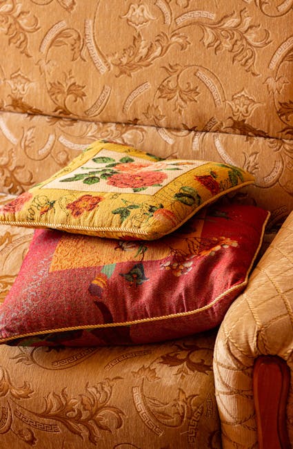 A living room scene featuring a blue upholstered sofa with a cream-colored throw blanket draped over its armrest, situated against a grey wall with a large green leafy plant nearby. Next to the sofa is a small, round, metallic side table with a polished rose gold finish, holding yellow rubber gloves, a pink microfiber cloth, and a transparent spray bottle filled with cleaning solution. The room is well-lit, highlighting the clean and tidy appearance of the furniture and decor. This image illustrates surface cleaning and domestic hygiene practices, aligning with Kingston Carpet Cleaners' upholstery cleaning and care services as seen on their Kingston Hill guide webpage.