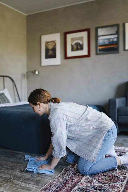 A person dressed in full protective white overalls, a face mask, and blue gloves is engaged in deep surface cleaning inside a residential living room. They are using a black vacuum hose to clean a lig